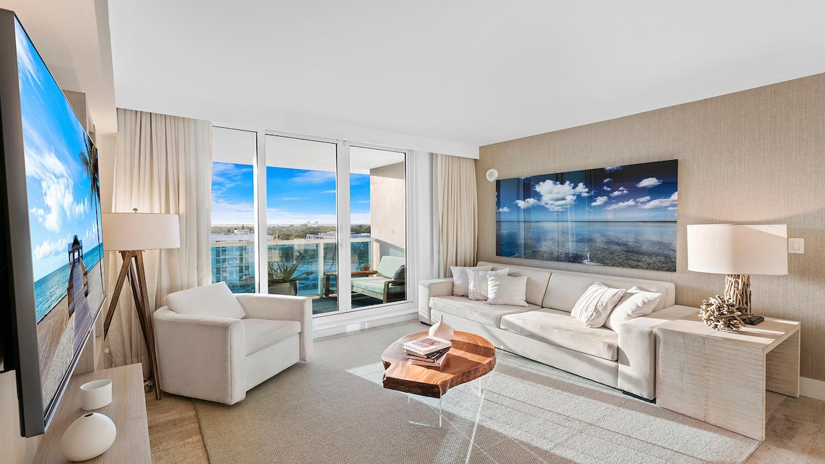 A spacious living room featuring a large flat-screen TV and a contemporary sofa set. Natural light is allowed in through expansive glass doors leading to a balcony, showcasing views of the city and bay. Light-colored walls and soft furnishings create a calming environment.