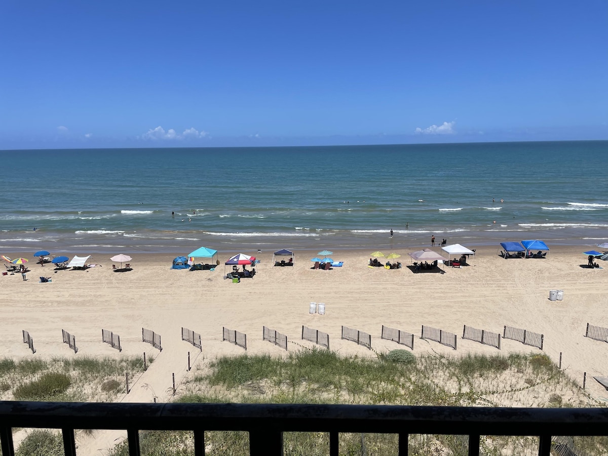 A wide sandy beach extends before the water, with several colorful umbrellas and beach chairs arranged along the shore. The gentle waves of the Gulf of Mexico reflect the clear blue sky, creating a serene coastal view perfect for relaxation.