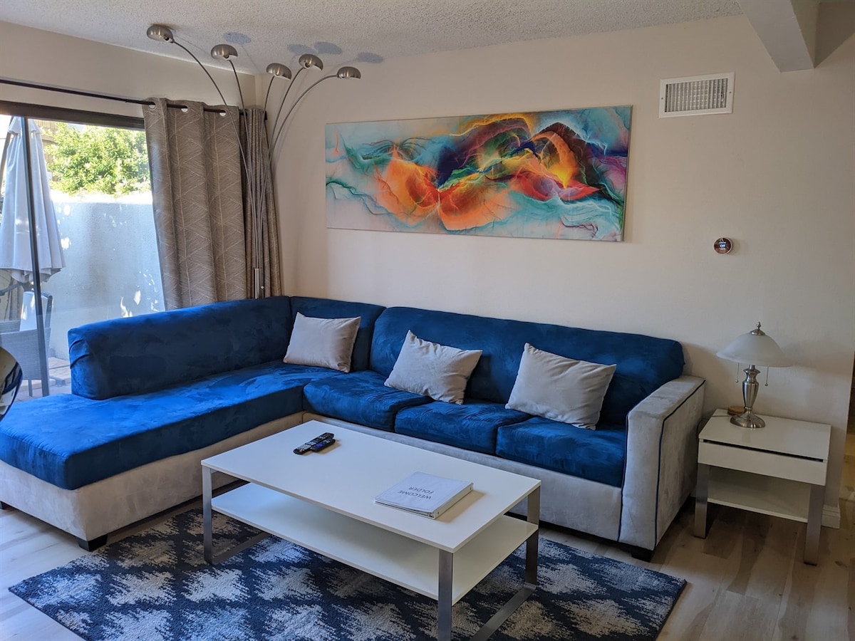 A modern living area features a large sectional sofa in blue with decorative cushions. A sleek coffee table is centered on a patterned area rug. Natural light enters through large sliding glass doors, partially covered by light curtains, enhancing the space's bright atmosphere.