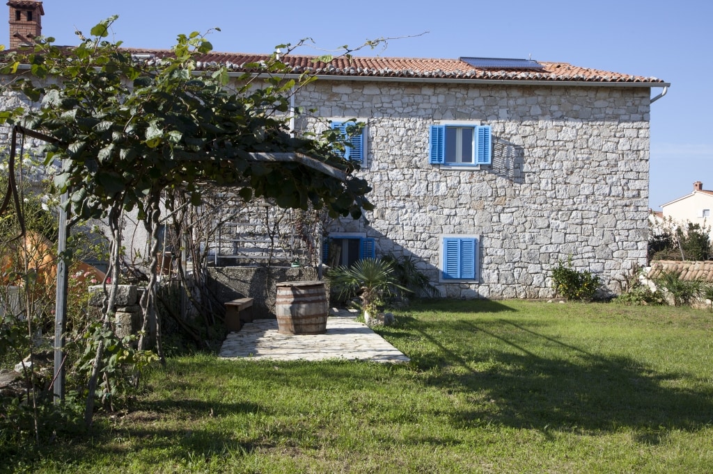 The exterior of the stone building is highlighted, showcasing blue shutters against the rustic facade. A spacious grassy area is visible in front, complemented by a wooden barrel and vine-covered pergola, creating a relaxed outdoor setting.