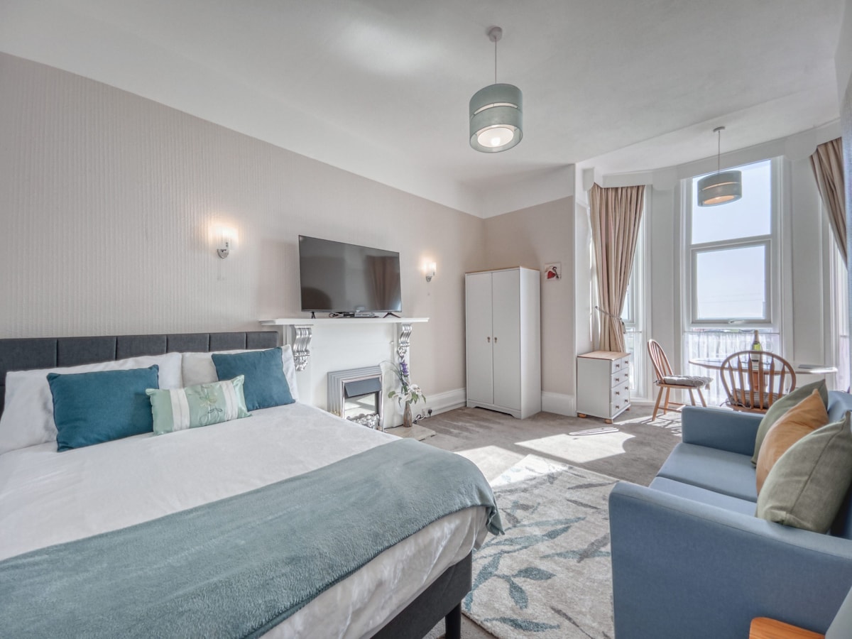 A spacious studio apartment features a comfortable sleeping area with a double bed dressed in soft linens. A seating area includes a sofa and a wooden chair, while large windows frame the sea view, inviting natural light into the room.