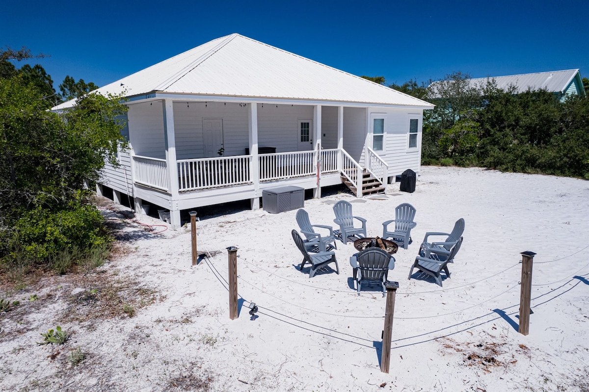 A spacious white house with a metal roof is surrounded by sandy terrain and greenery. The exterior features a wide porch with steps leading up, while a circular arrangement of six Adirondack chairs encircles a wood-burning fire pit, creating a cozy outdoor gathering space.