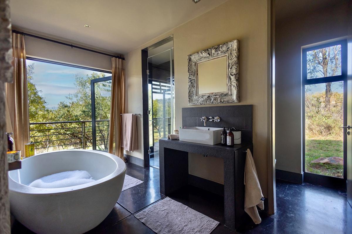 A spacious bathroom features an oval stone bathtub and a modern wash basin set on a dark granite countertop. Large windows provide views of the bushveld, while a separate glass shower opens to the outdoors. Neutral tones and soft textiles enhance the serene environment.