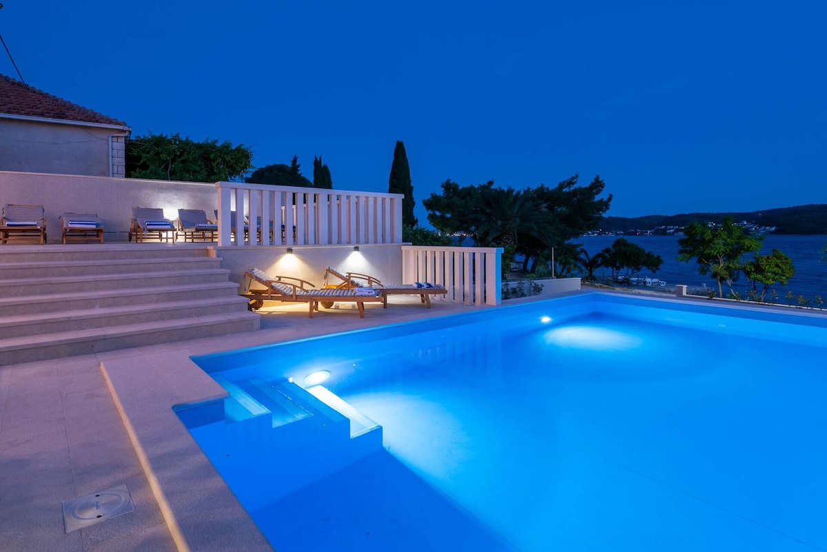 An inviting swimming pool is captured under the evening sky, illuminated in soft blue tones. Comfortable loungers are arranged nearby, with steps leading from the pool area. Lush greenery and the shimmering water of the nearby channel complete the serene atmosphere.