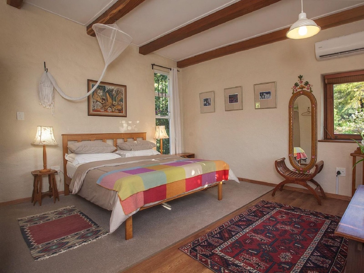A cozy sleeping area features a spacious bed adorned with a colorful blanket. A wooden framed mirror stands against the wall, complemented by two bedside lamps that provide soft lighting. Natural light filters in through the window, adding warmth to the room.