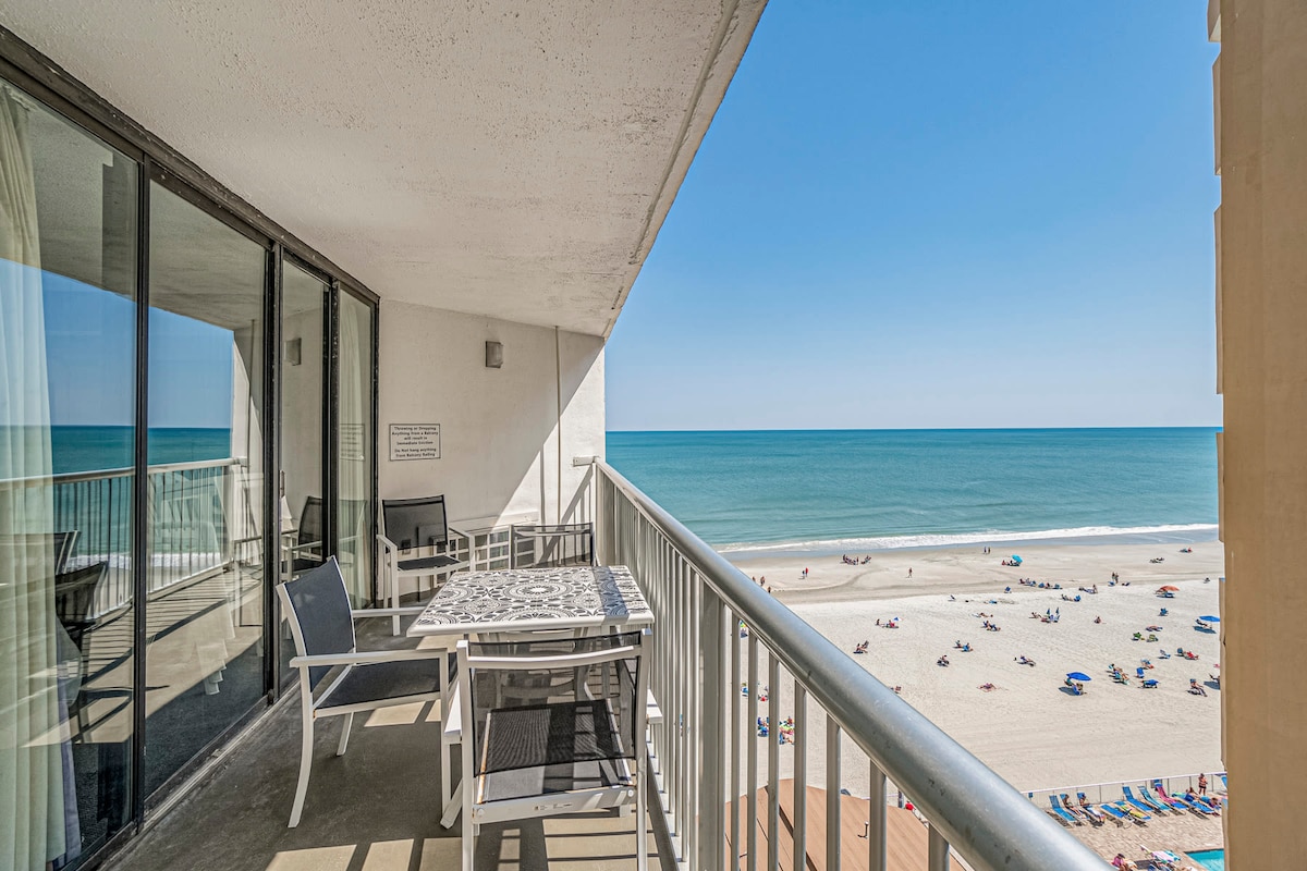 A spacious balcony overlooks the Atlantic Ocean, with a table and chairs positioned for enjoying views of the beach. The ocean's calm waters stretch out to the horizon, while sunbathers and colorful umbrellas are visible on the sandy shore below.