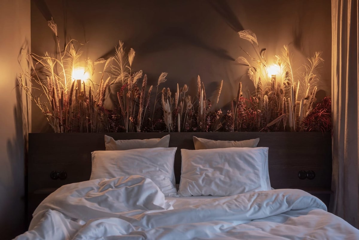 A spacious bed is positioned against a dark wall, complemented by a floral art installation of dried flowers above the headboard. Soft lighting from two sconces enhances the cozy ambiance. Wrinkled white bedding suggests a welcoming and restful environment.