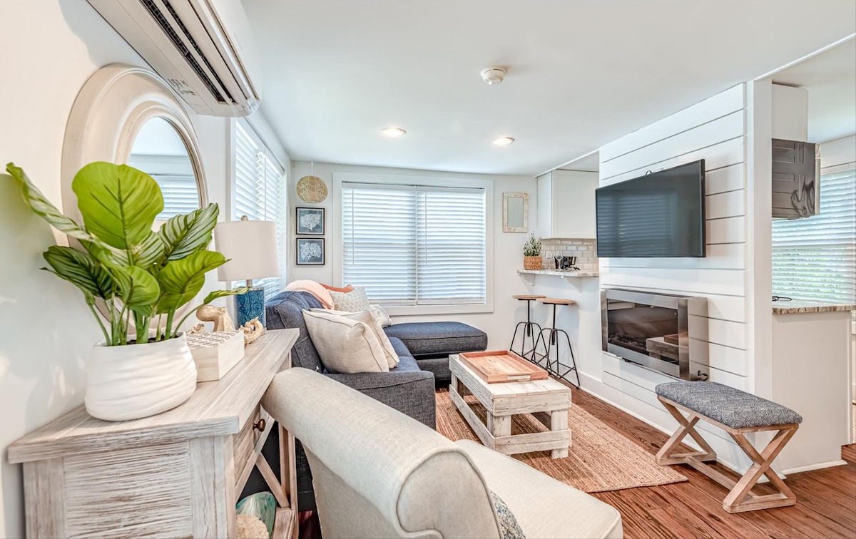 A cozy living area is highlighted by a navy blue sofa and a light-colored armchair, arranged around a wooden coffee table. Natural light filters through the window with blinds. Decor includes green plants and a wall-mounted TV. A small dining area is visible in the background.