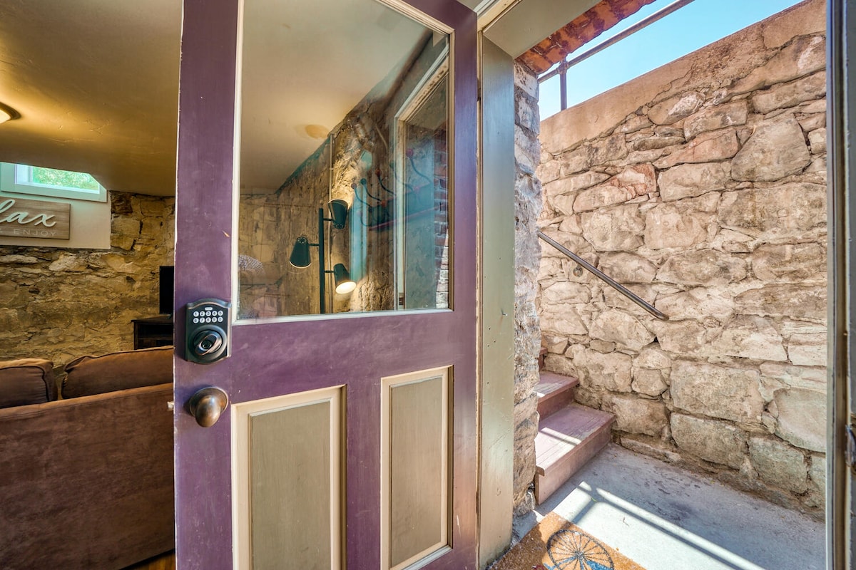 A private entry door features a keypad lock and opens to a well-lit space with unique stone walls. Natural light filters in through a small window at the top of the stairs, leading to the cozy living area. Steps descend to the main part of the home.