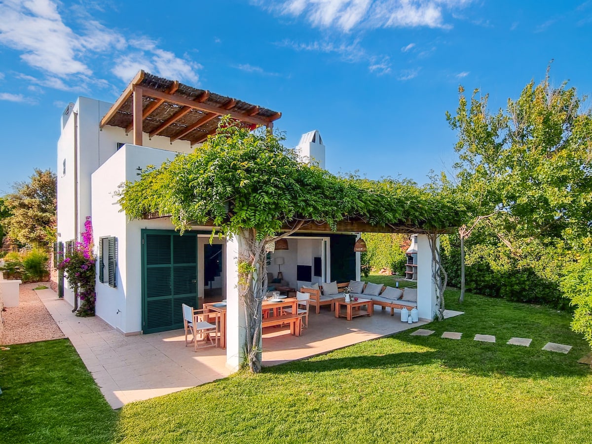 A spacious outdoor area is surrounded by lush greenery, featuring a covered patio with comfortable seating and a dining arrangement. The white villa is complemented by vibrant plants, with a glimpse of the upper terrace visible. A well-maintained lawn extends beyond the seating area, inviting relaxation.