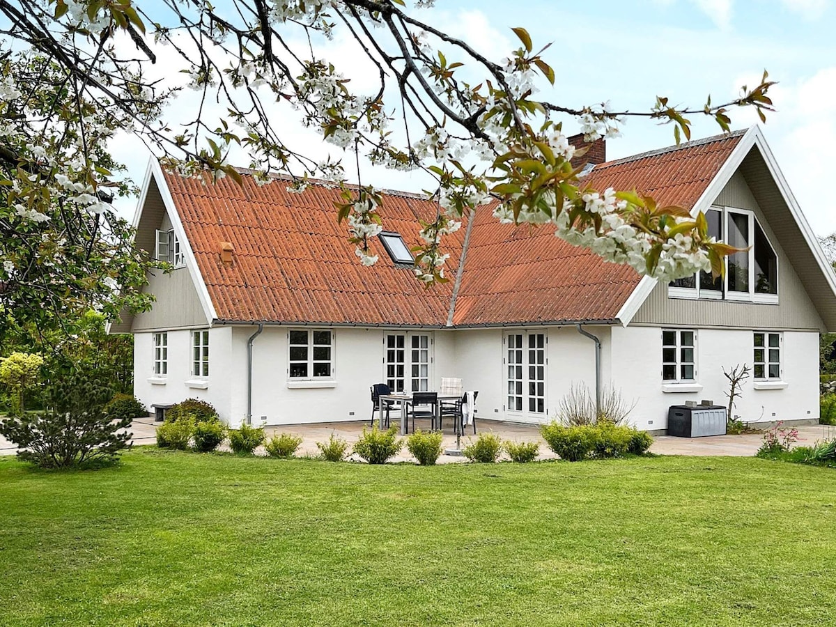 A spacious holiday home is set within a lush green lawn, surrounded by neatly trimmed hedges and flowering plants. The house features a distinctive red-tiled roof and large windows, providing a bright and inviting exterior. A patio area with outdoor furniture is visible for relaxation.