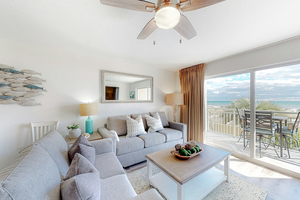 A living area is furnished with a large sectional sofa and accent chairs, complementing a coffee table adorned with decorative items. Natural light flows through the sliding glass doors, providing a view of the ocean and beach. A ceiling fan and lamps add to the comfortable ambiance.