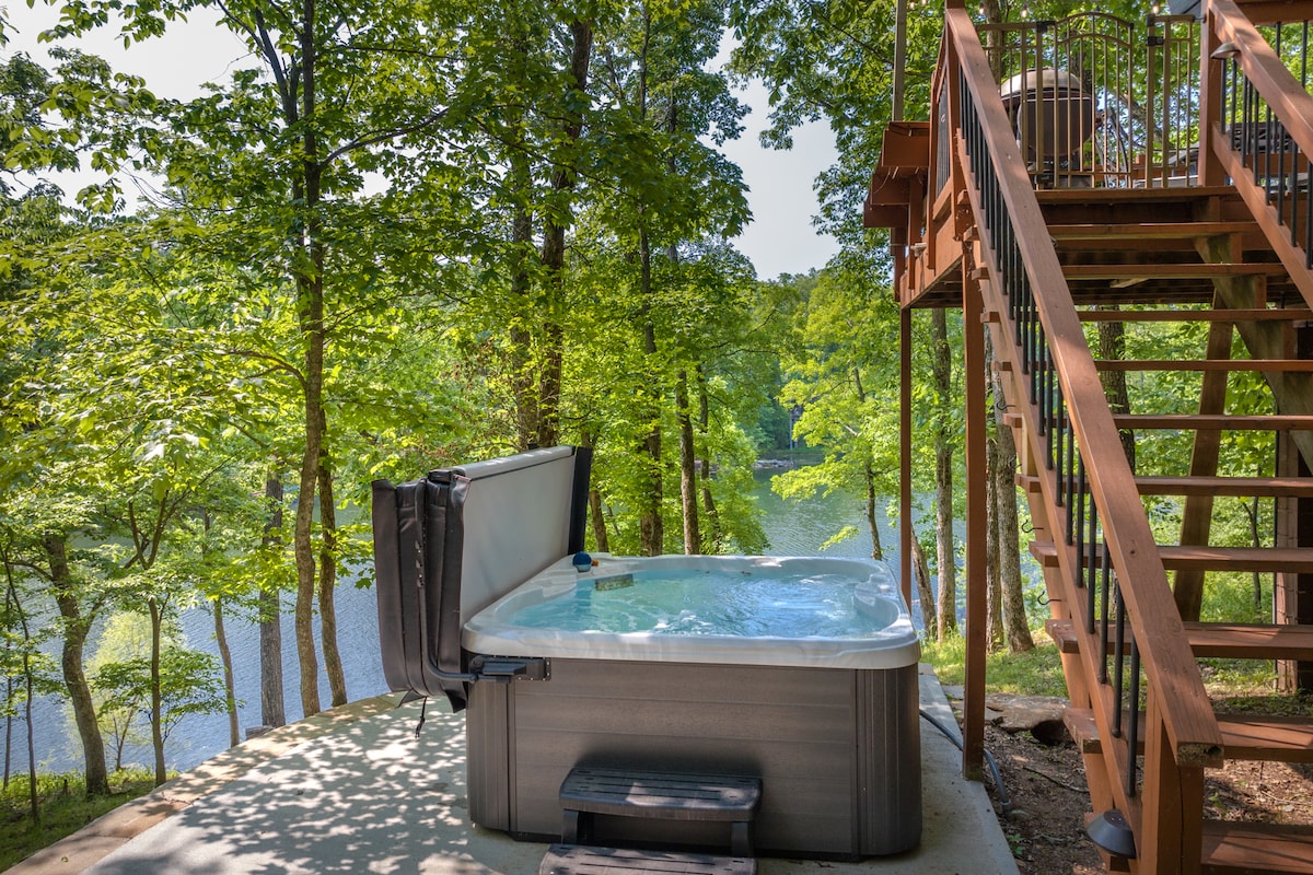 A hot tub is situated on a patio surrounded by lush greenery, providing a serene view of the lake. Steps lead up to a deck area, enhancing the outdoor experience amidst the natural setting.