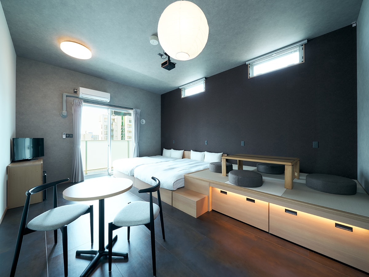 A spacious room features two beds arranged neatly, complemented by a low table and cushioned seating on a raised platform. Natural light enters through large windows, enhancing the modern design with neutral tones and minimalist furnishings.