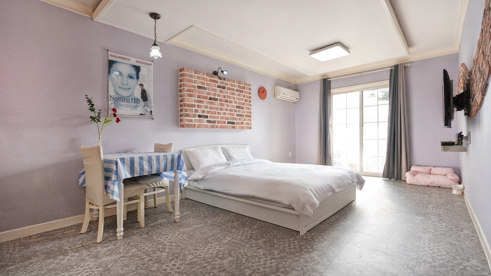 The spacious room features a large bed with soft bedding and a light-colored duvet. A dining table with a blue and white checkered tablecloth sits beside the bed. Natural light enters through a glass door leading to a small outdoor area.