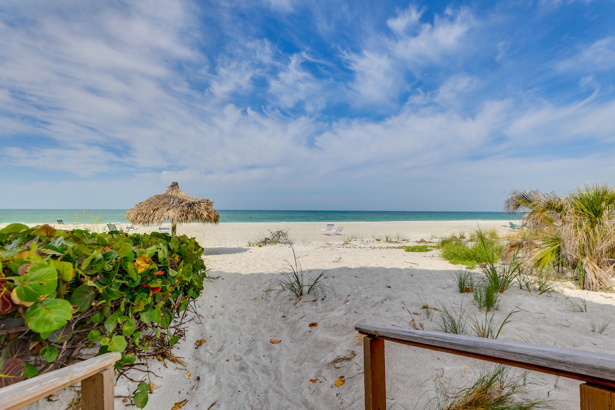 A sandy path leads to a private white-sand beach, with a thatched umbrella visible near the shoreline. The calm turquoise water stretches towards the horizon under a partly cloudy sky, creating a serene coastal atmosphere.