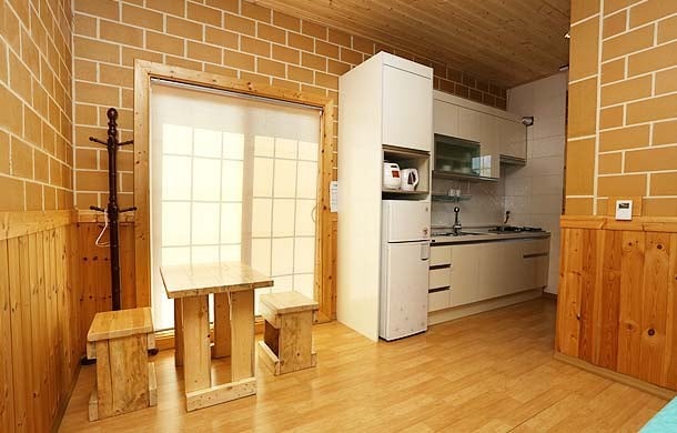A simple kitchen area features wooden walls and flooring, complemented by a dining table and two benches crafted from the same wood. A refrigerator and stove are present in the kitchenette, with natural light streaming through a large window adorned with a sheer curtain.