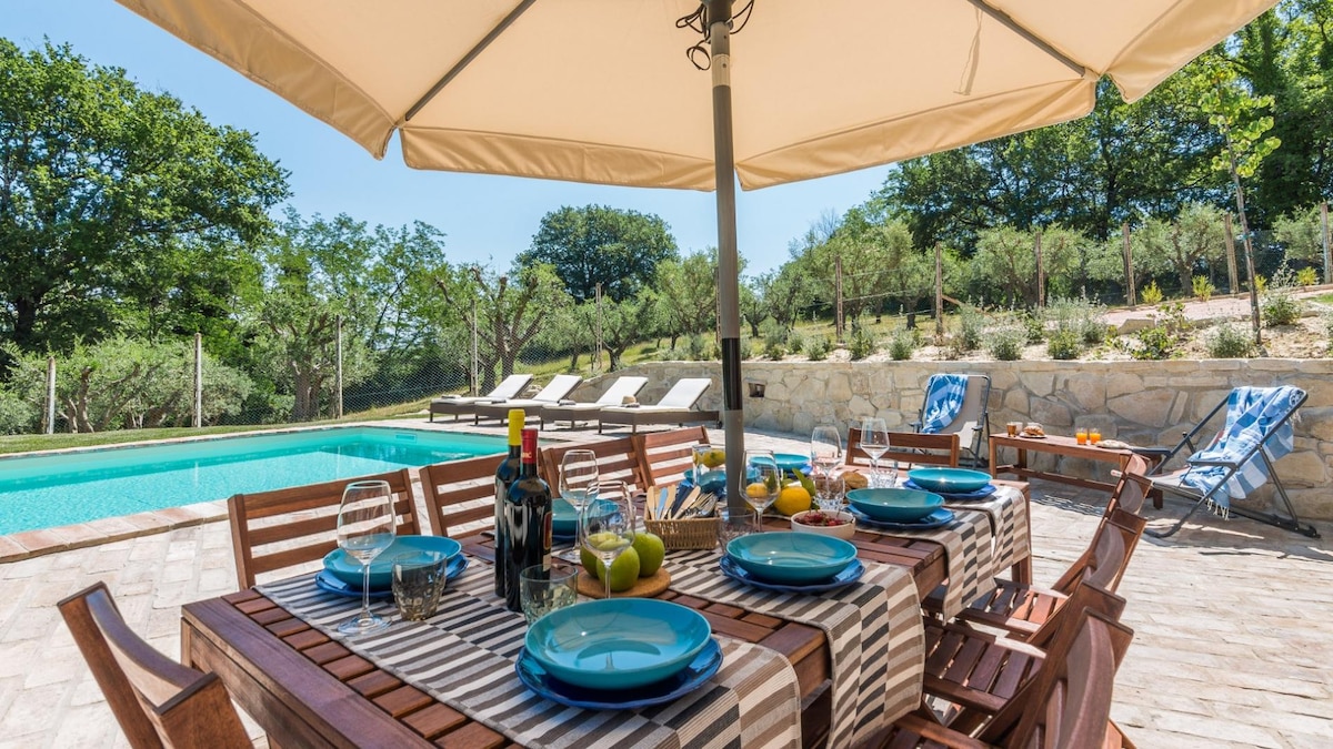 An outdoor dining area is set beneath a large umbrella, featuring a wooden table adorned with blue plates and crystal glasses. A basket holds fresh fruit. The swimming pool glistens nearby, surrounded by lounge chairs and a backdrop of greenery.
