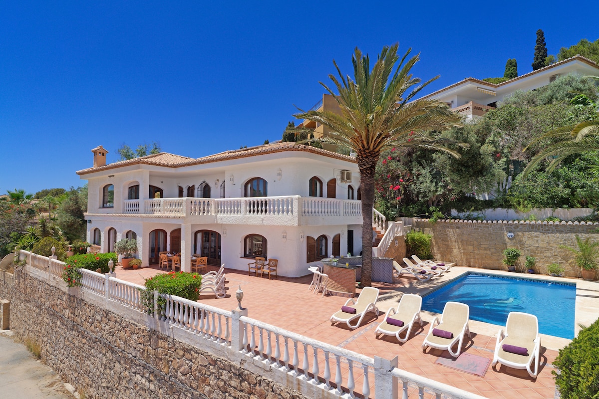 The villa presents a spacious exterior with a welcoming façade featuring multiple balconies and large windows. A solar-heated pool is surrounded by sun loungers, and palm trees provide shade in the sunny landscape. The terracotta terrace is designed for relaxation and enjoyment.