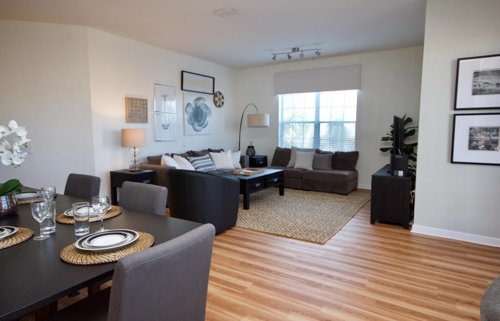 An open-plan living and dining area is presented, featuring a dark wood dining table set for six and a comfortable seating arrangement with a large sectional sofa. Natural light floods the space, enhanced by light-colored walls and wood flooring, creating a welcoming environment.