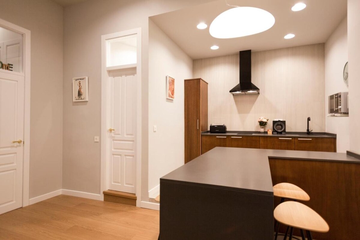 A modern kitchen area is featured with a dark countertop and wooden cabinetry. Two light-colored stools sit at the breakfast bar. An extractor hood is installed above the stove, and appliances, including a microwave, are neatly arranged on the countertop. Bright ceiling lights illuminate the space.