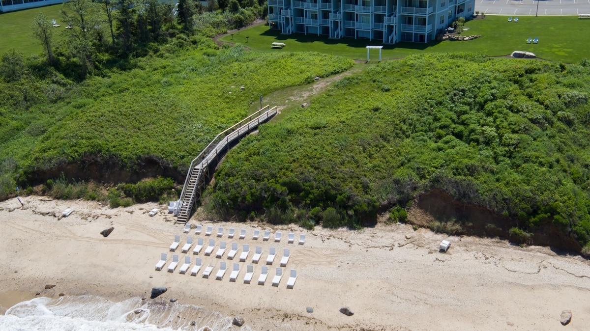 A sandy beach area is visible, lined with white lounge chairs arranged in neat rows. A wooden walkway leads from the green hillside to the beach. A low-rise building is positioned in the background, surrounded by lush greenery.