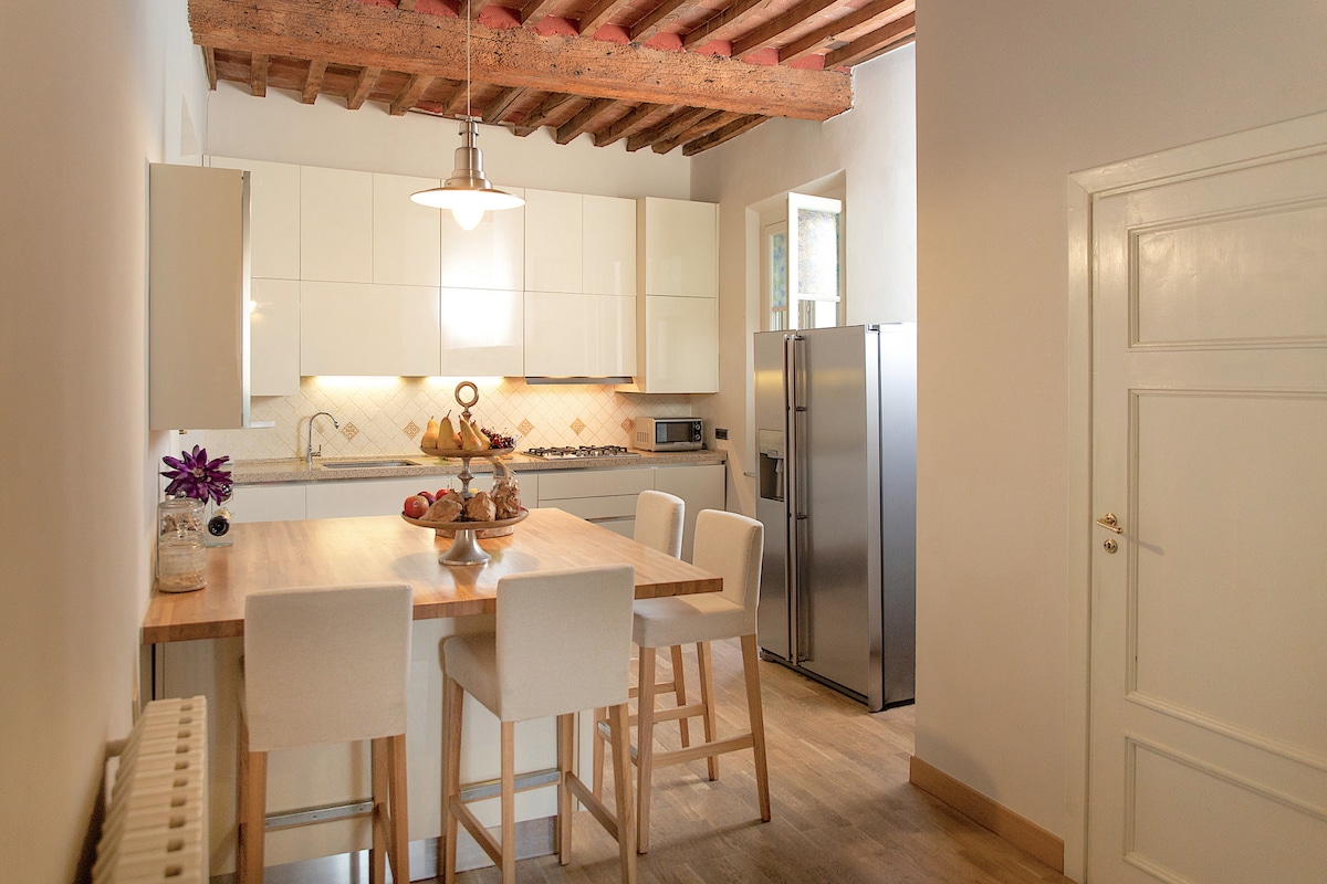 A modern kitchen features a light wood dining table surrounded by four upholstered chairs. The space is well-lit, showcasing white cabinetry and stainless steel appliances. Exposed wooden beams add a touch of character, while a window offers natural light.