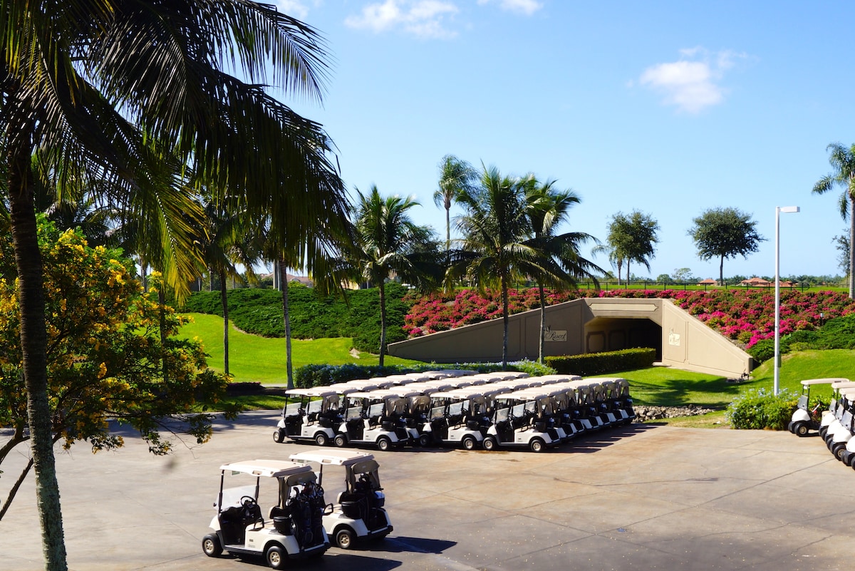 A parking area is showcased, filled with numerous white golf carts lined up orderly. Lush green lawns and flowering bushes with vibrant colors surround the entrance to a clubhouse, providing a tropical backdrop under a clear blue sky.