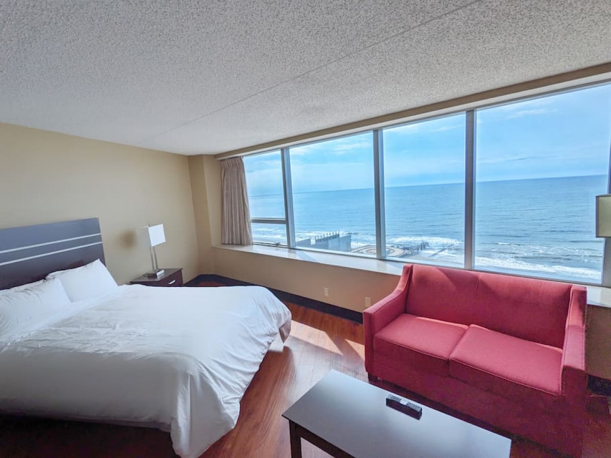 A spacious bedroom features a large bed dressed in white linens, complemented by a red couch positioned near floor-to-ceiling windows. The ocean view stretches beyond the beach, reflecting natural light into the room. A bedside table and lamp enhance the functionality of the space.