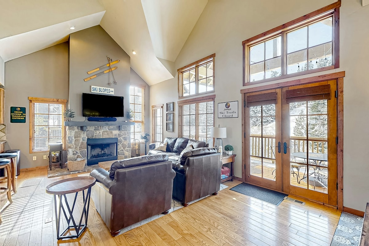 A spacious living area is highlighted by large windows that allow natural light to fill the room. Leather sofas are arranged around a stone fireplace, with a TV mounted above. Double doors lead to an outdoor deck, creating a seamless connection to the outside.