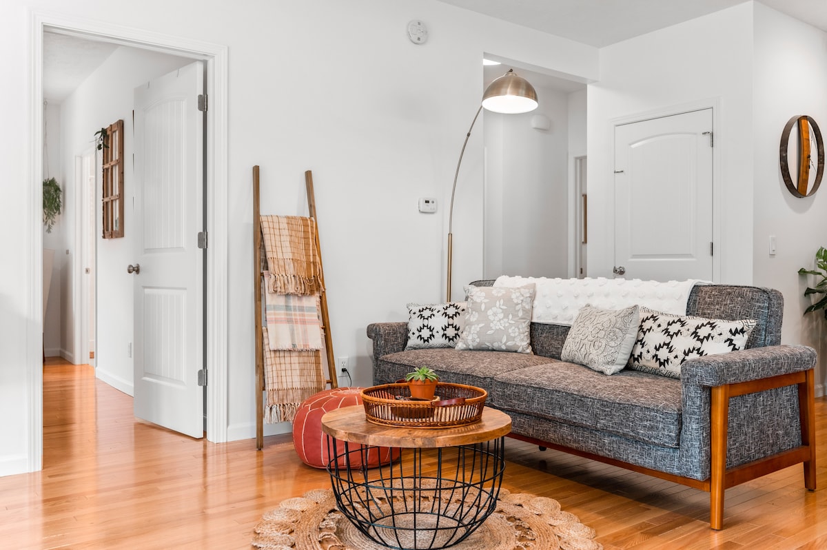 The living room features a modern sofa adorned with patterned cushions, a round coffee table nestled on a textured rug, and warm wooden flooring. The space is complemented by natural light streaming in from nearby windows and a floor lamp providing additional illumination.
