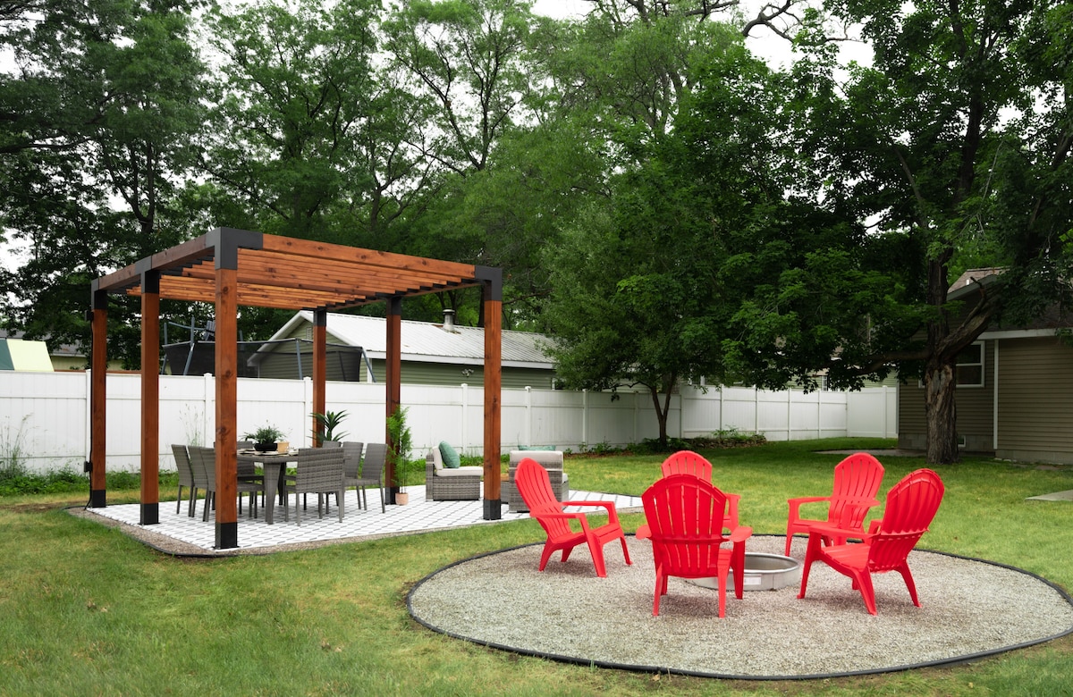 A spacious backyard features a wooden pergola providing shade over a dining area, furnished with a table and chairs. Bright red Adirondack chairs surround a gravel fire pit, creating an inviting space for outdoor relaxation and gatherings.