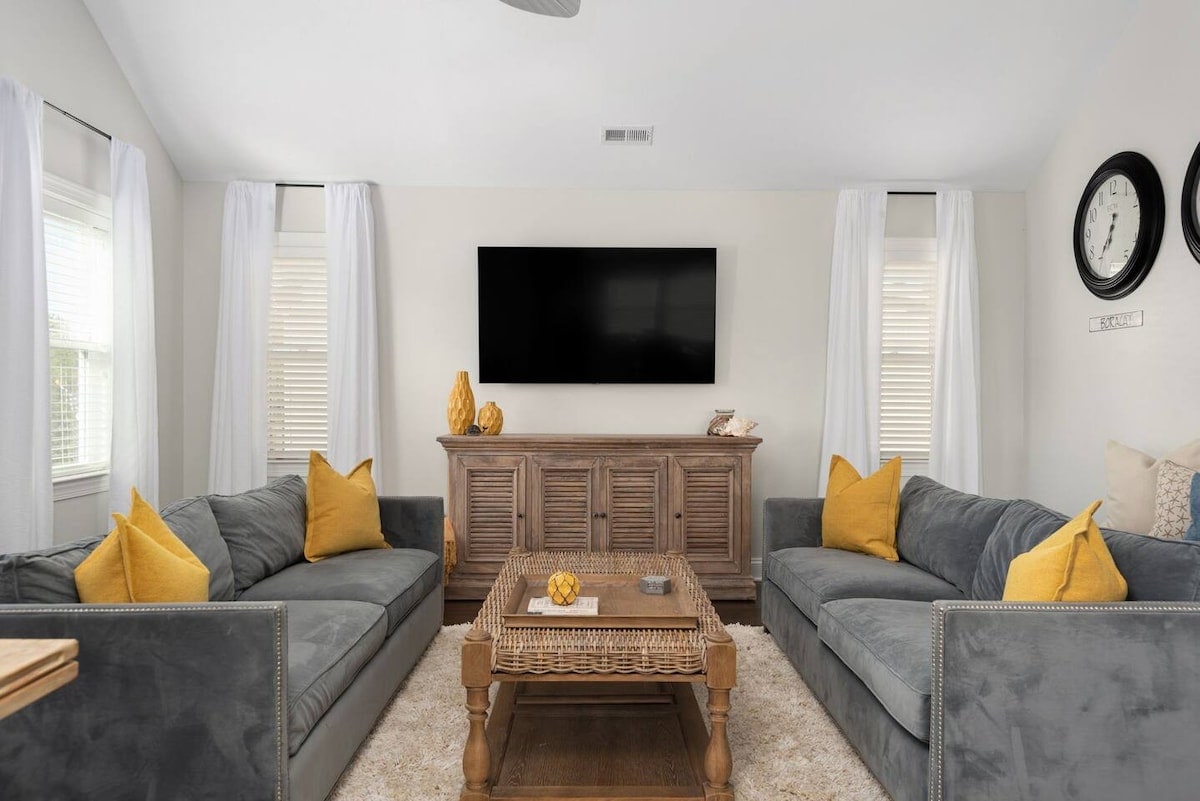 A spacious living room features two plush sofas with yellow accent pillows, framed by large windows covered with sheer curtains. A natural wood coffee table sits in the center, complemented by a decorative bowl. A flat-screen TV is mounted on the wall above a wooden media cabinet.