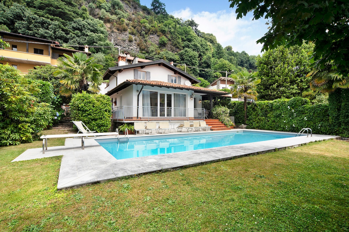 A spacious villa is surrounded by lush greenery and features a clear swimming pool. Sun loungers are placed along the poolside, which is framed by a manicured lawn. The house offers a covered terrace area with steps leading to the pool.