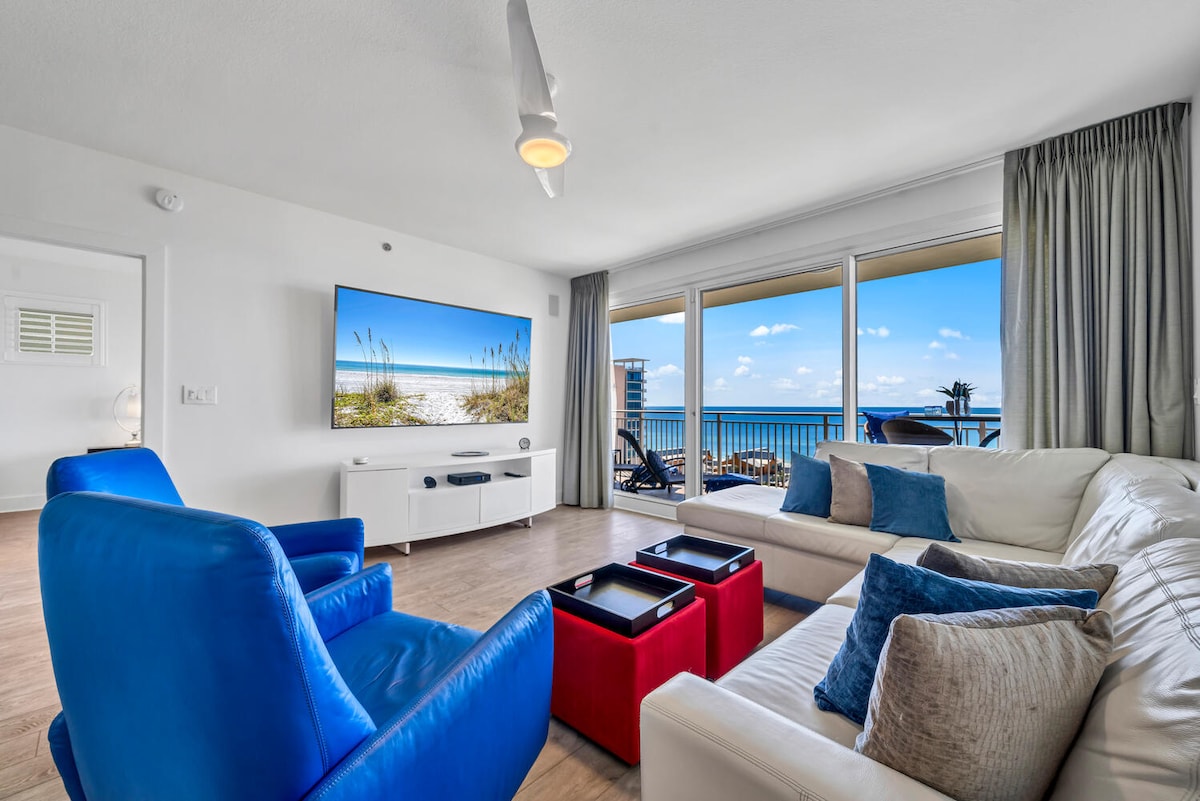 A spacious living area features a modern white sectional sofa with decorative cushions, facing a wall-mounted television. Large glass doors frame a view of the Gulf, enhancing natural light. Vibrant blue chairs add color to the neutral decor, creating a welcoming ambiance.