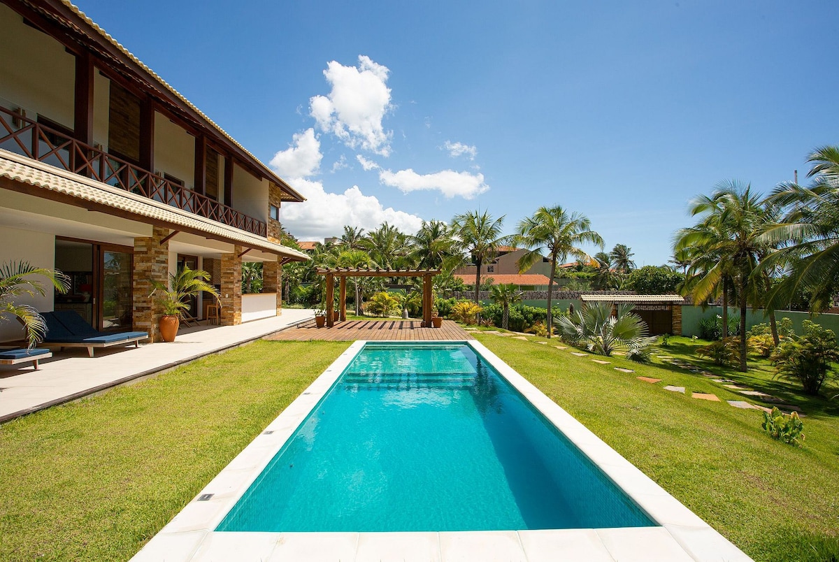 An outdoor area showcases a refreshing swimming pool surrounded by well-maintained gardens and palm trees. A wooden deck and dining space are located nearby, providing a serene setting. The villa's two-story architecture is visible in the background under a bright blue sky.