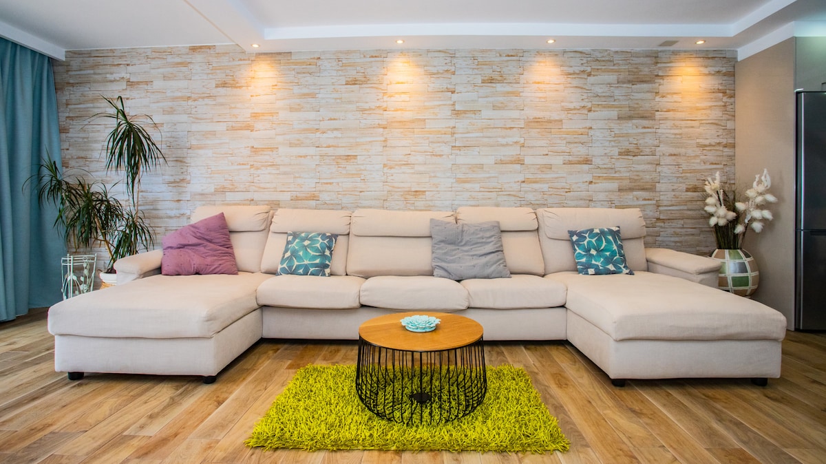 A spacious living area features a light-colored sectional sofa adorned with decorative pillows in various shades. A round coffee table with a patterned top sits on a bright green area rug, creating a cozy gathering space. Soft lighting highlights a textured stone wall backdrop.