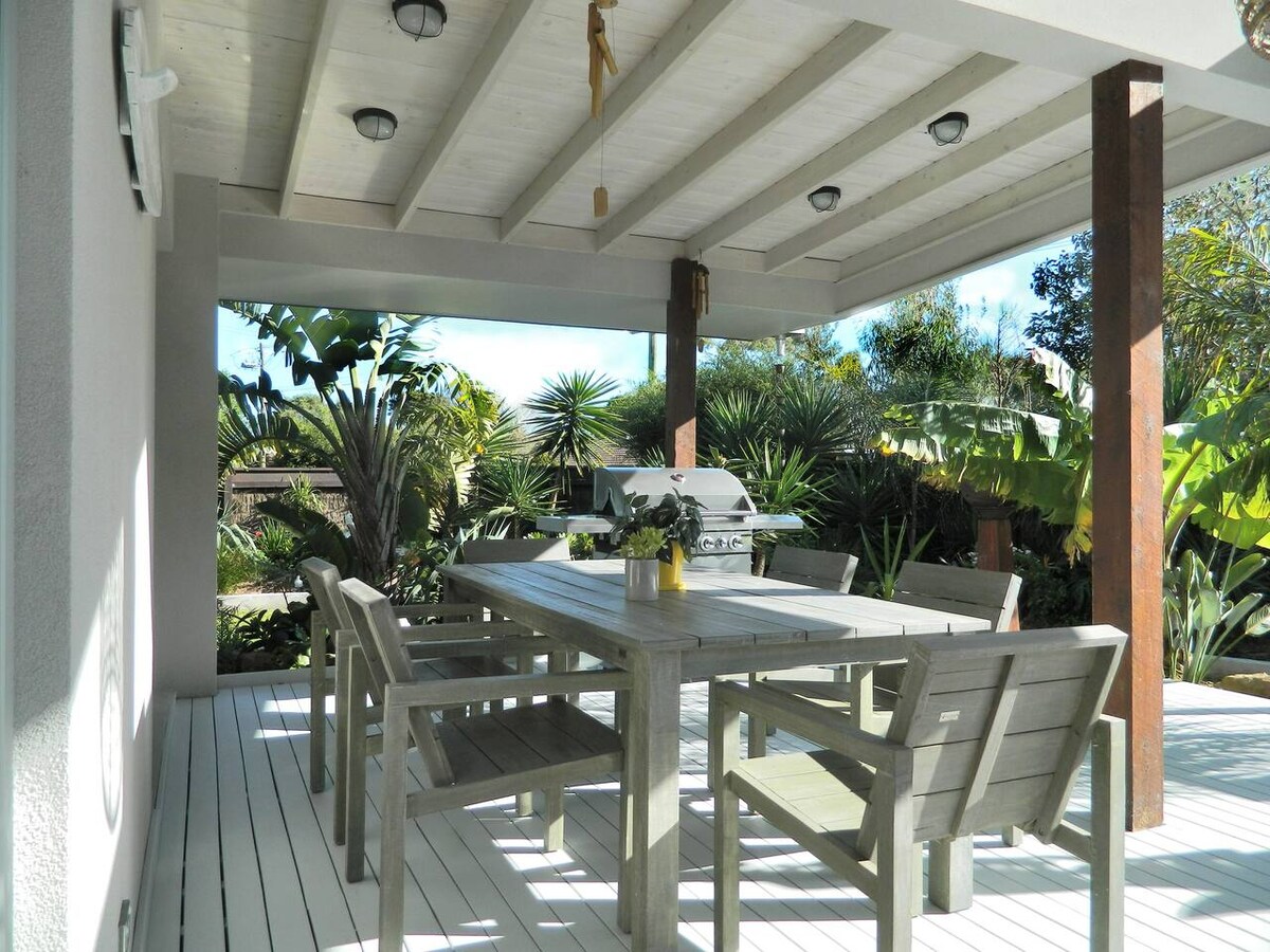 An alfresco dining area features a large wooden table surrounded by multiple chairs under a covered patio. Lush greenery and tropical plants create a serene environment, while overhead lights add warmth to the space.