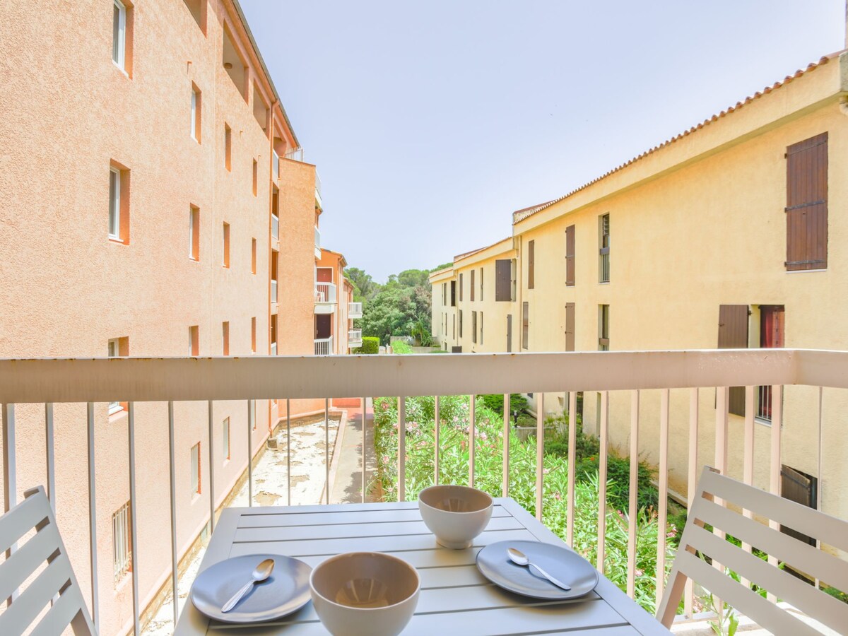A balcony showcases a white table set with two neutral bowls and cutlery. Surrounding views of neighboring buildings and greenery are visible, providing a sense of openness in a quiet courtyard setting.