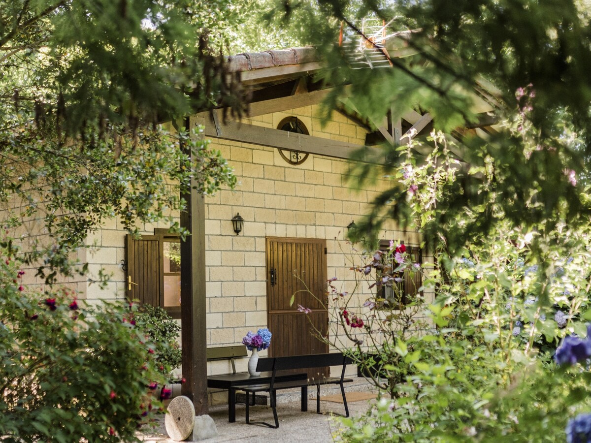 The exterior of the casita is framed by lush greenery, featuring a smooth stone façade and wooden doors. A simple bench is positioned in front, accompanied by colorful flowers, creating a charming and serene environment.