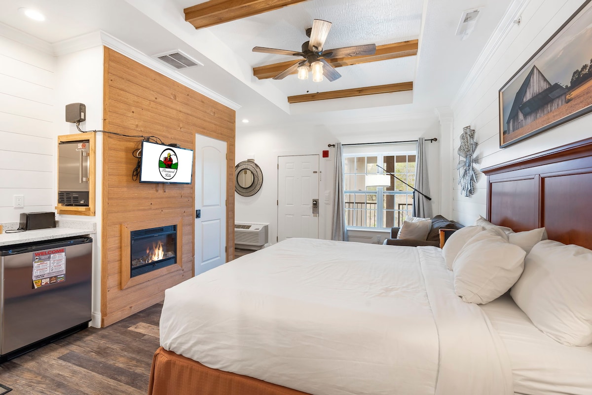A queen suite features light wood accents and a spacious bed with crisp white linens. A modern kitchenette with appliances is situated nearby, and a wall-mounted TV is visible. Access to a balcony with natural light is provided through sliding doors, enhancing the room's comfort.