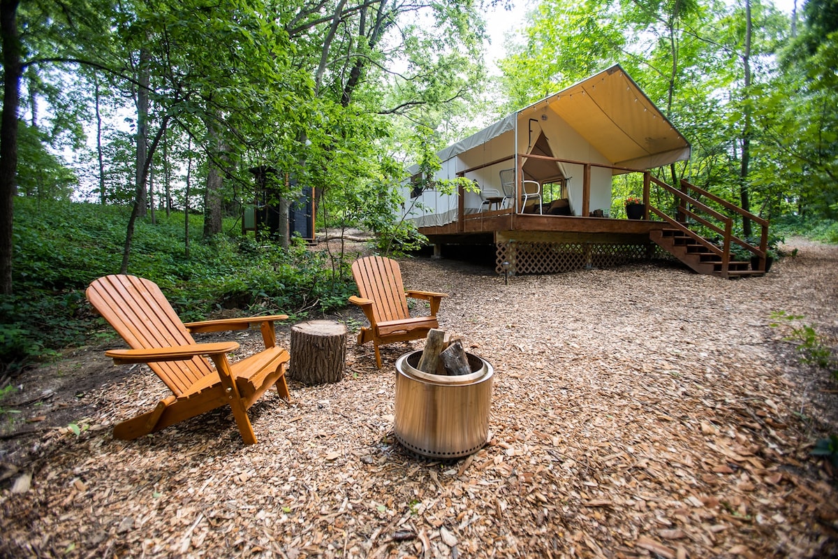 A glamping tent is nestled among trees, featuring a wooden deck with steps leading up to the entrance. Two Adirondack chairs face a fire pit, surrounded by gravel and tree bark, creating a cozy outdoor space for relaxation.
