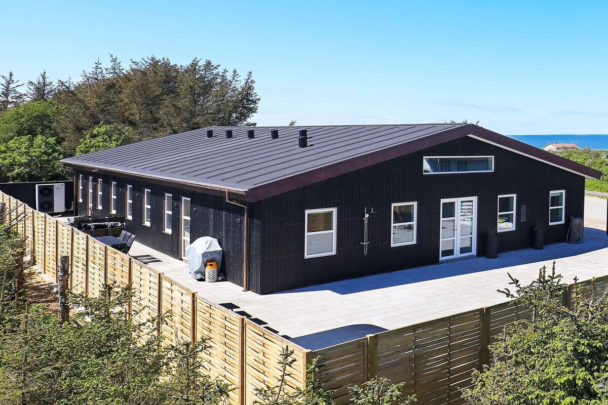 The exterior of the holiday home is showcased, featuring a contemporary black wooden design. A stone patio is visible, surrounded by a wooden fence. Nearby, outdoor amenities are positioned, including a barbecue grill. The ocean can be glimpsed in the background, framed by lush greenery.