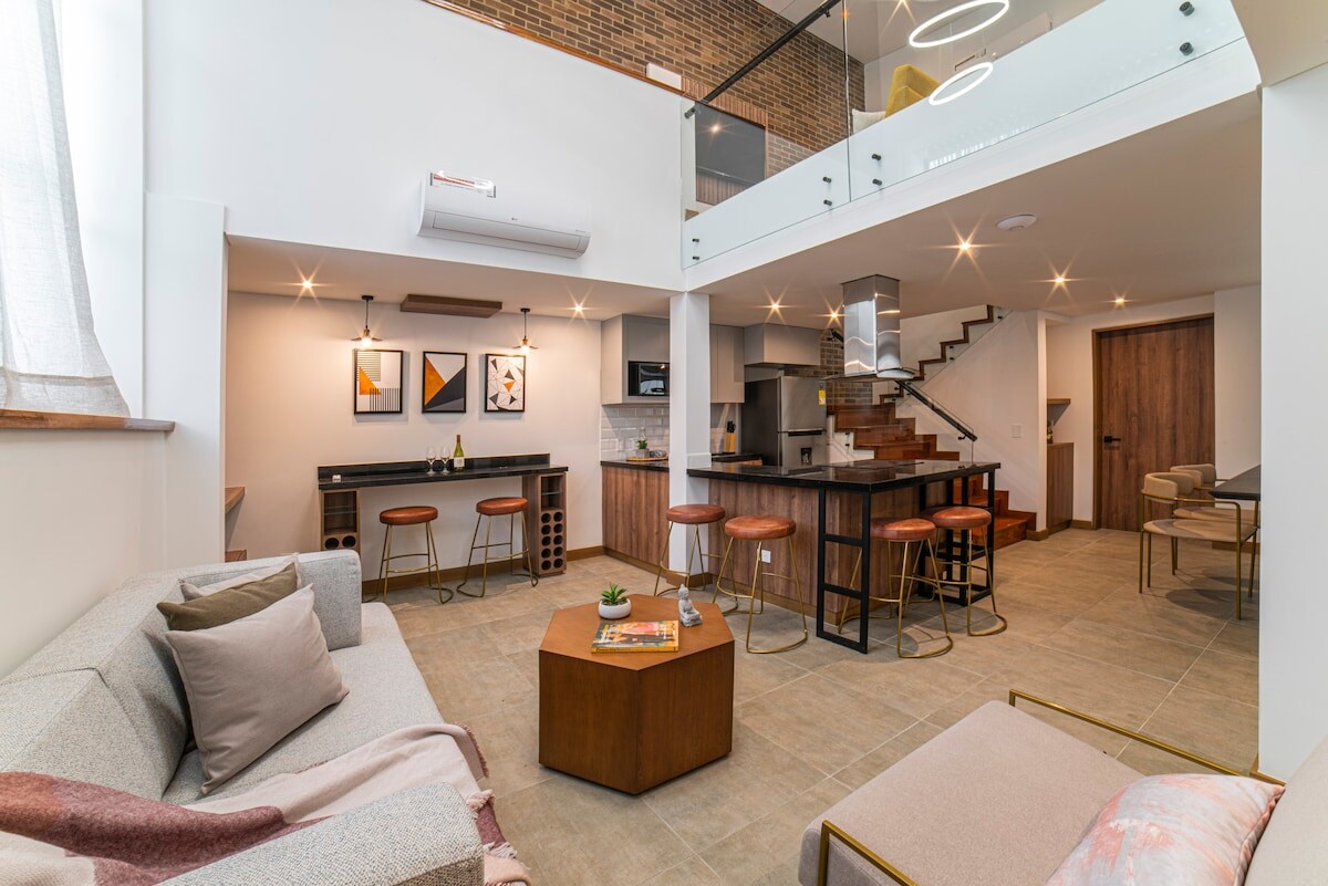 The living area showcases an open layout with a contemporary design. Soft seating is complemented by a wooden coffee table, while bar-style seating at the kitchen island offers additional dining options. Natural light floods the space through large windows, enhancing the inviting atmosphere.