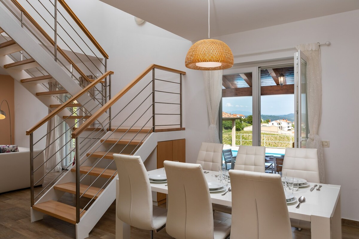 A modern dining area is shown with a white table set for six, surrounded by cushioned white chairs. Natural light enters through large windows, revealing a scenic view. A wooden staircase leads to the upper floor, enhancing the spacious atmosphere.