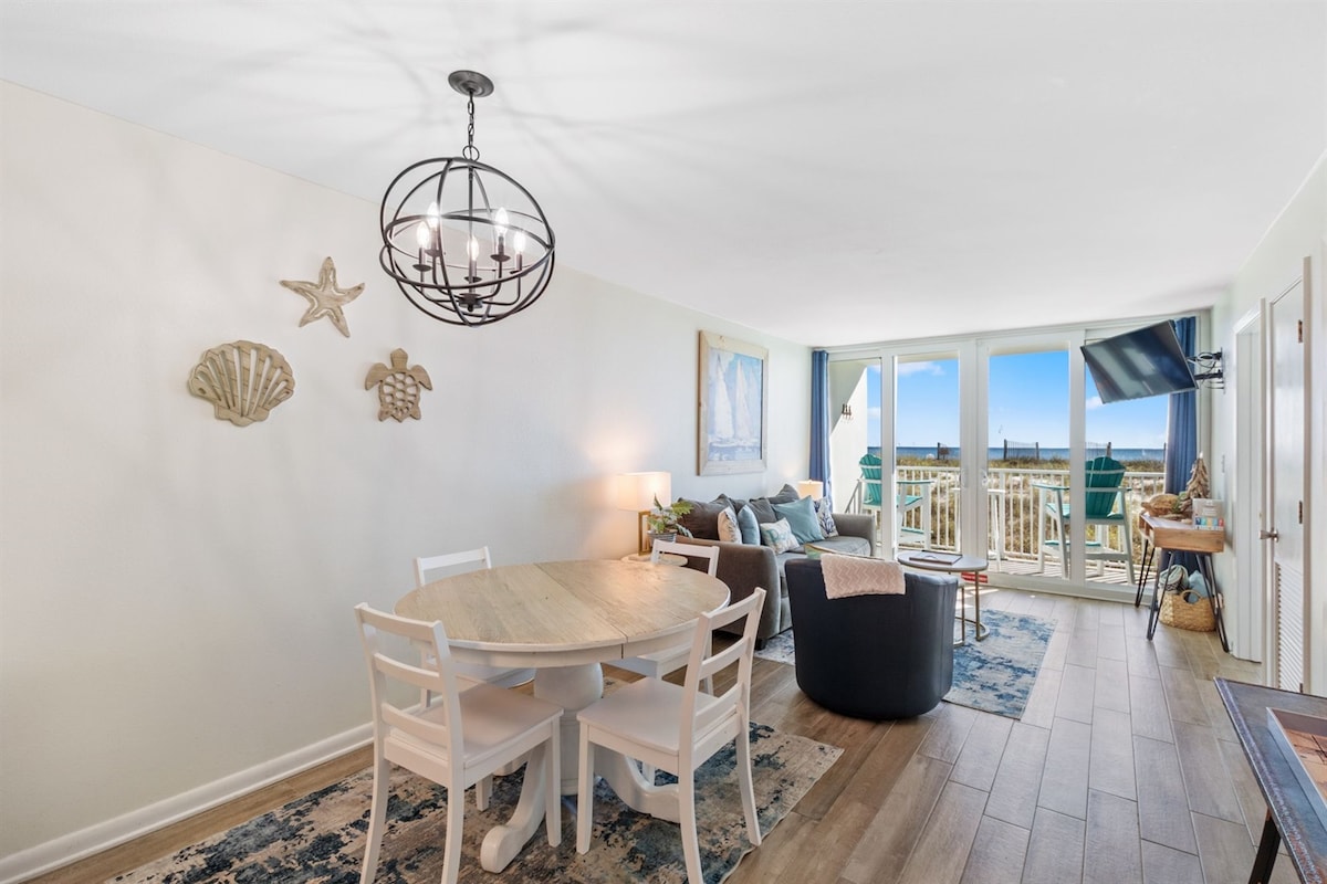 A bright living area is furnished with a round dining table and four white chairs. Adjacent, a cozy seating arrangement includes a dark armchair and a sofa, with decorative pillows. Large glass doors allow natural light and offer views of the beachfront, complemented by coastal-themed decor.