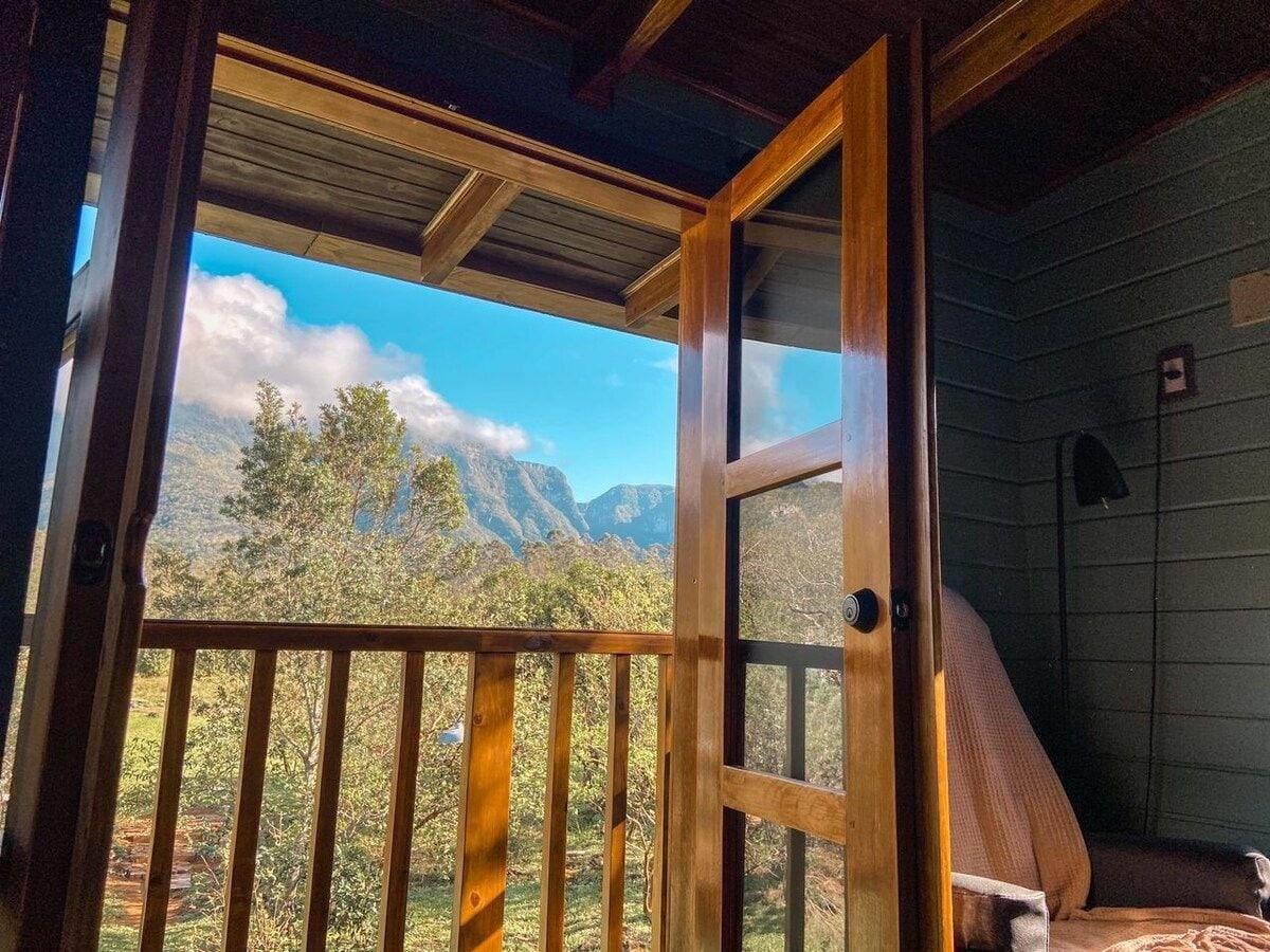 A balcony door with wooden frames opens to reveal a panoramic view of the mountainous landscape. Lush greenery and distant hills are visible, along with a serene blue sky. A comfortable armchair is positioned nearby, inviting relaxation.