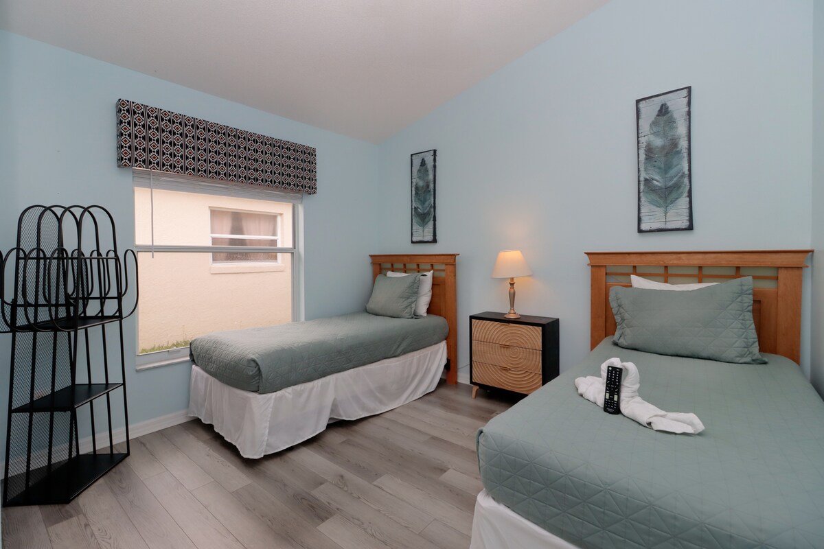 Привлекателна спалня с две легла в Кисими, близо до Дисни Орландо, с меко зелено спално бельо и произведения на изкуството от пера – идеална за спокойна нощ.