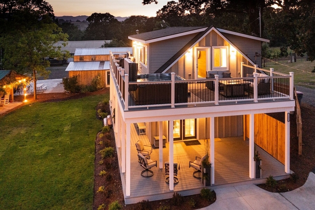 A spacious two-story home is nestled in a green landscape, featuring a large covered porch and outdoor seating. The upper deck showcases a hot tub, surrounded by railings. Soft lights illuminate the exterior, creating a cozy atmosphere at twilight.