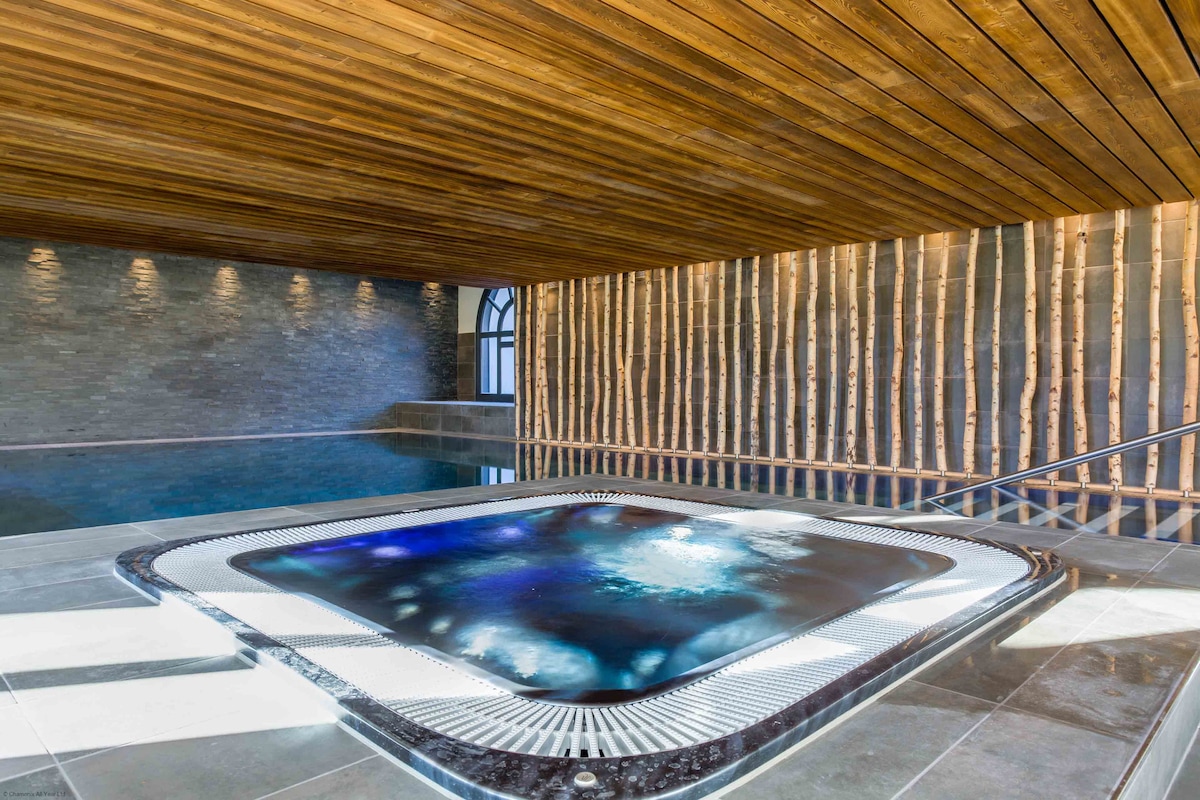 The indoor spa area features a heated swimming pool alongside a jacuzzi, with modern tile and wood finishes. Soft lighting enhances the natural textures of the wooden ceiling and stone walls, creating a calming atmosphere.